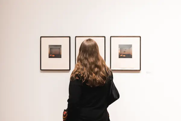 A woman with light brown hair looking at 3 paintings hanging on the wall at Opening Reception of the exhibition Subjective Illuminations, featuring prints from the collection - image courtesy of Juicebox Studios