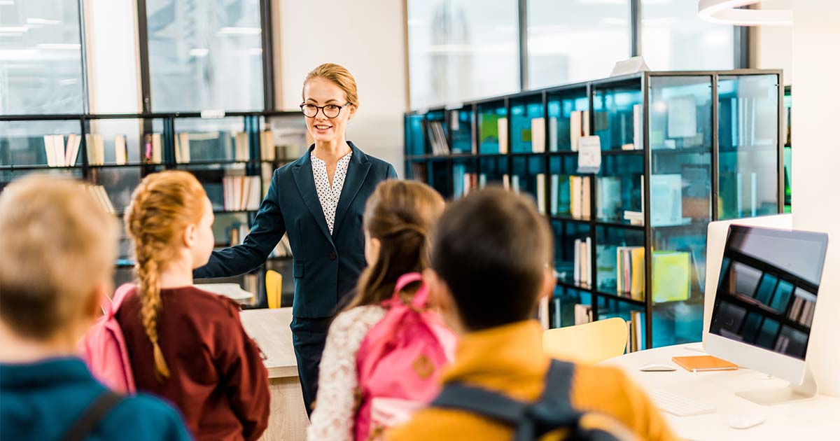 3 Ways to Lead from Within the School Library post cover image: A teacher in a library smiles and gestures while speaking to a group of attentive students with backpacks. Bookshelves and computers are in the background.