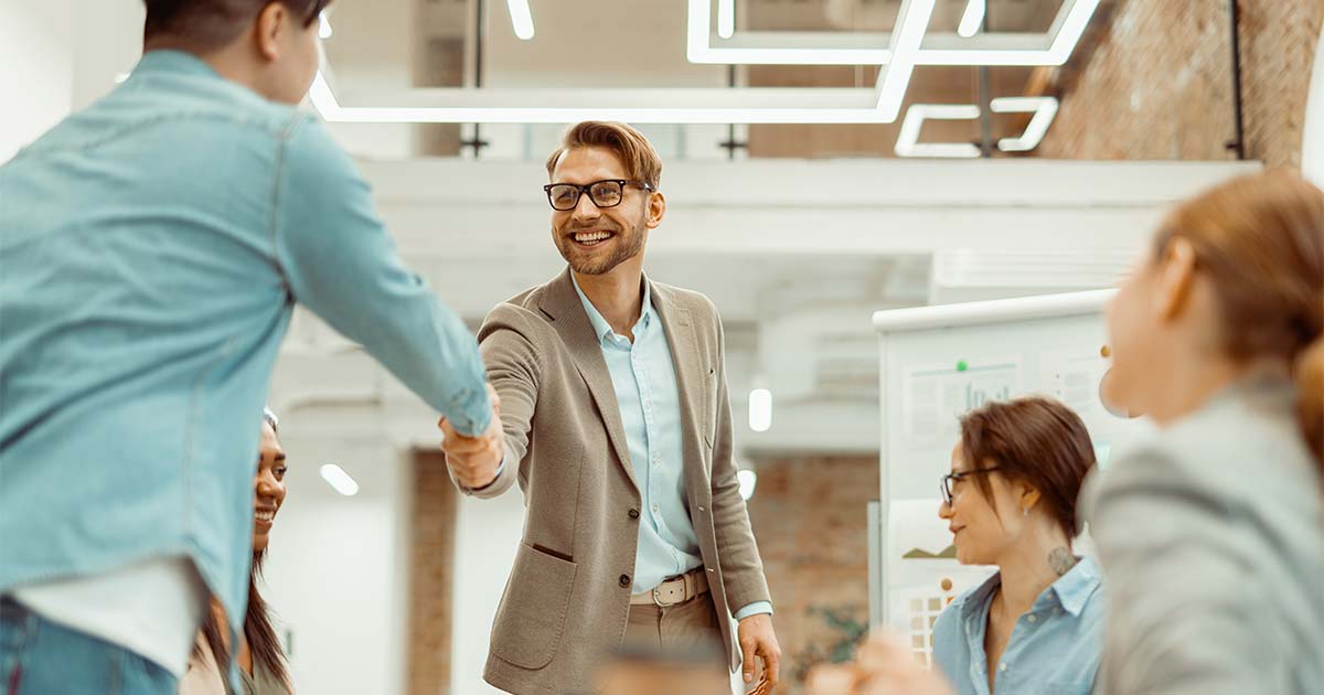 Building Partnerships for Archival Impact post cover image: Archivists meeting in a modern meeting space. A smiling man in a light colored blazer and khakis shakes hands with a casually dressed person. Colleagues, seated, listen around a table.