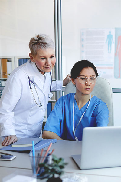Two healthcare professionals in an office setting. A woman in a white coat stands, smiling, while another in blue scrubs sits focused on a laptop that could be accessing Lucidea's Presto KM platform.