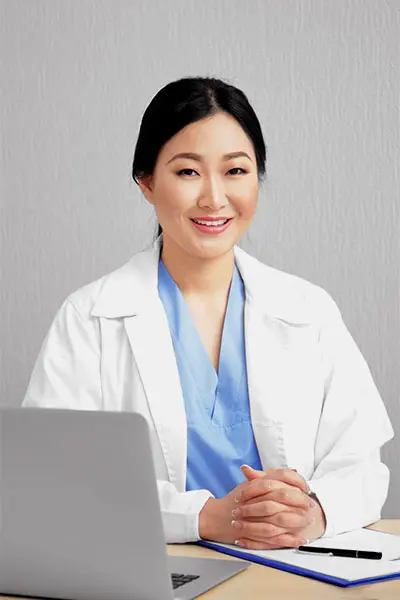 Asian female doctor in a white coat and blue scrubs sitting at a desk with a laptop and notebook, smiling warmly. The background is a textured gray wall.