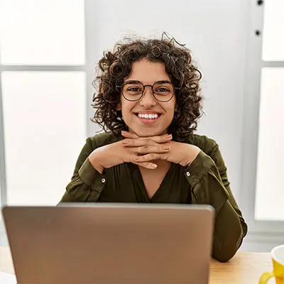 Smiling person with curly hair and glasses sits at a desk with a laptop. They rest their chin on their hands, conveying a friendly and welcoming tone.
