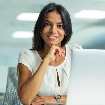 A woman in a white blouse sits at a desk with a laptop, smiling confidently after talking to Lucidea about Presto. The office setting is bright and modern, a professional atmosphere.