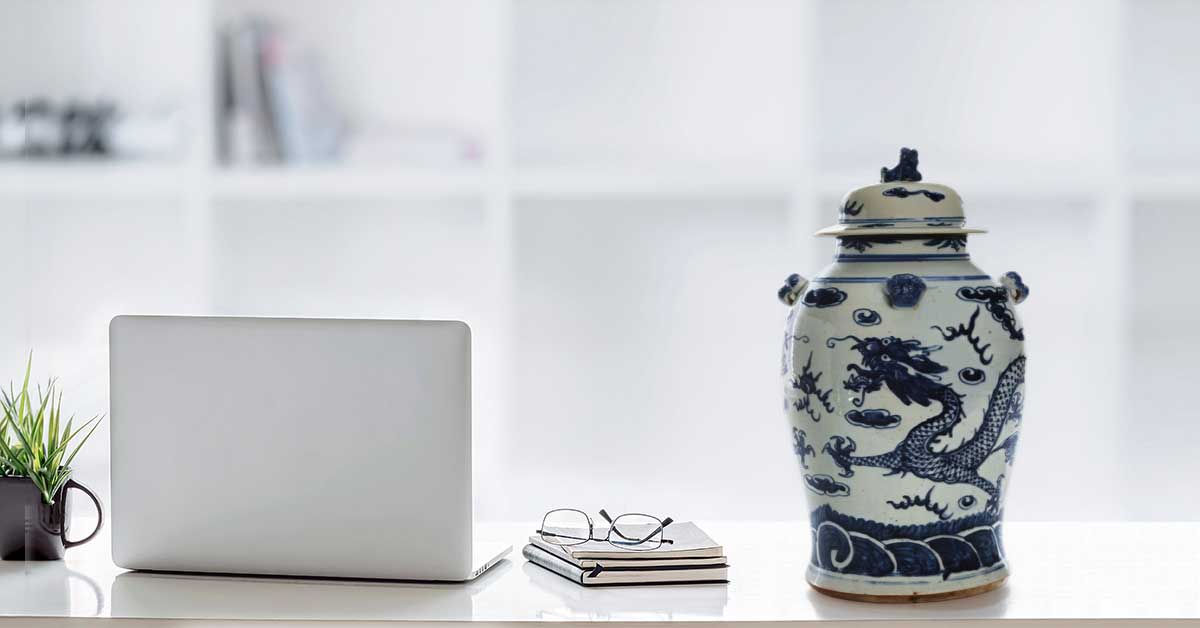 A laptop on a white desk beside a blue-and-white antique Chinese porcelain jar, with notebooks, reading glasses, and a small potted plant.