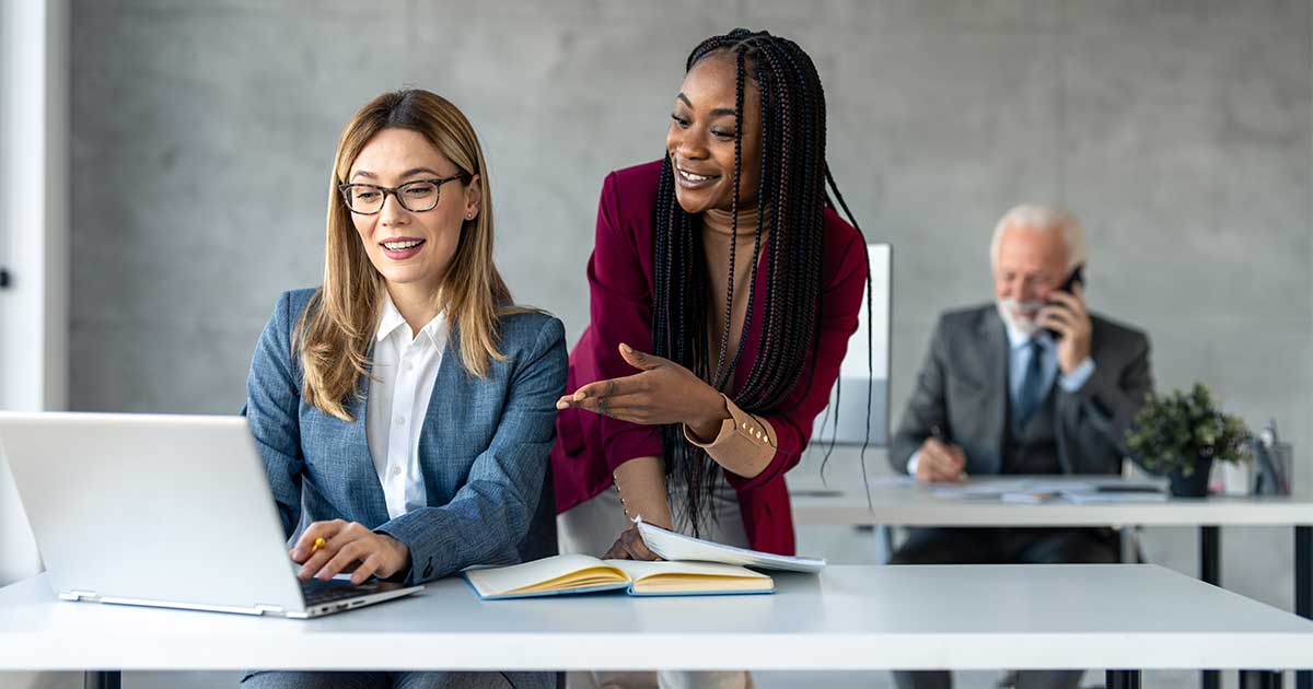 Cover image for the blog post, The KM Mentor, Part 5: The First 90 Days, by Stan Garfield featuring Two diverse businesswomen collaborate at a laptop and open notebook in a modern office, smiling and discussing Knowledge Management; an older man talks on the phone behind them.