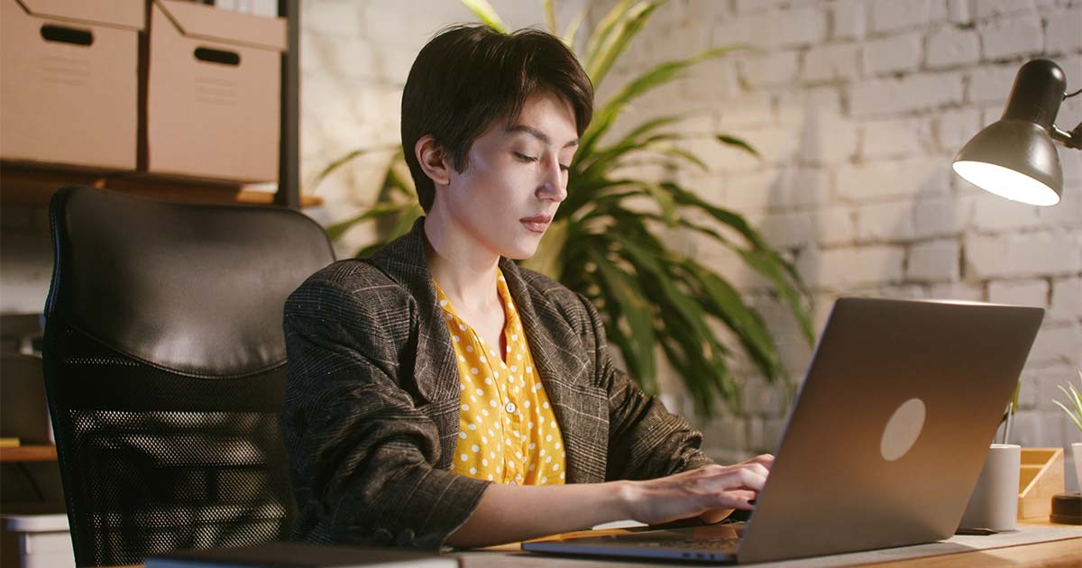 Cover image for Information Overload—How Special Librarians Can Stay Focused by Lauren Hays, PhD. featuring a focused professional woman typing on a laptop at a desk in a warm office, with a plant, desk lamp, brick wall, and storage boxes behind her.