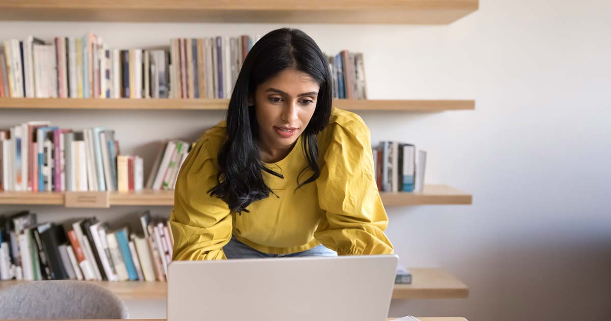 A librarian typing on a laptop in front of a wall of bookshelves.