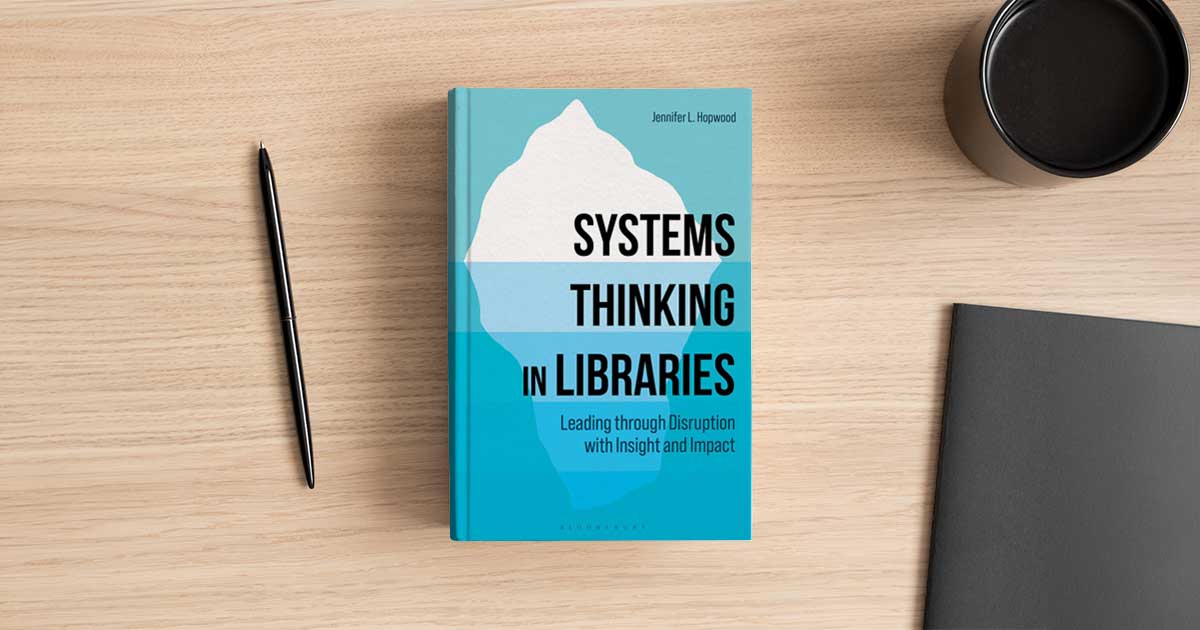 The book Systems Thinking in Libraries by Jennifer L. Hopwood, with an iceberg on its cover, on a wooden desk beside a pen and a coffee cup.