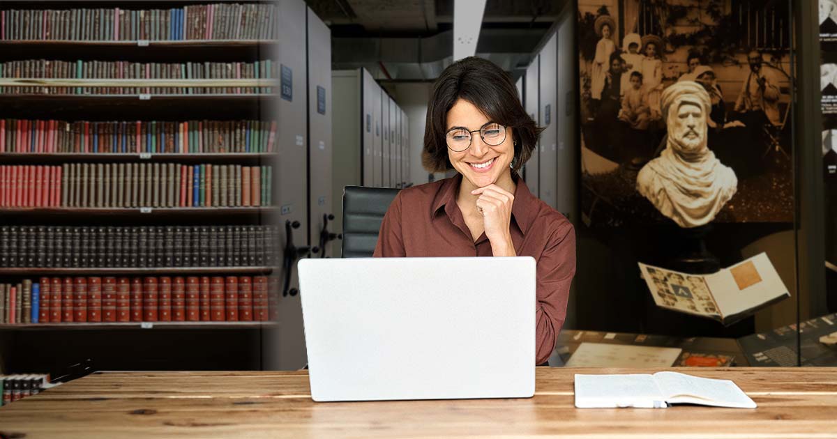 A person smiling at their laptop with bookshelves, archival storage, and a historical bust in the background.
