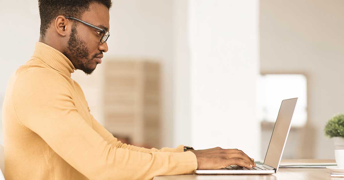 A man in glasses works on a laptop at a bright desk, representing online research and discovery.