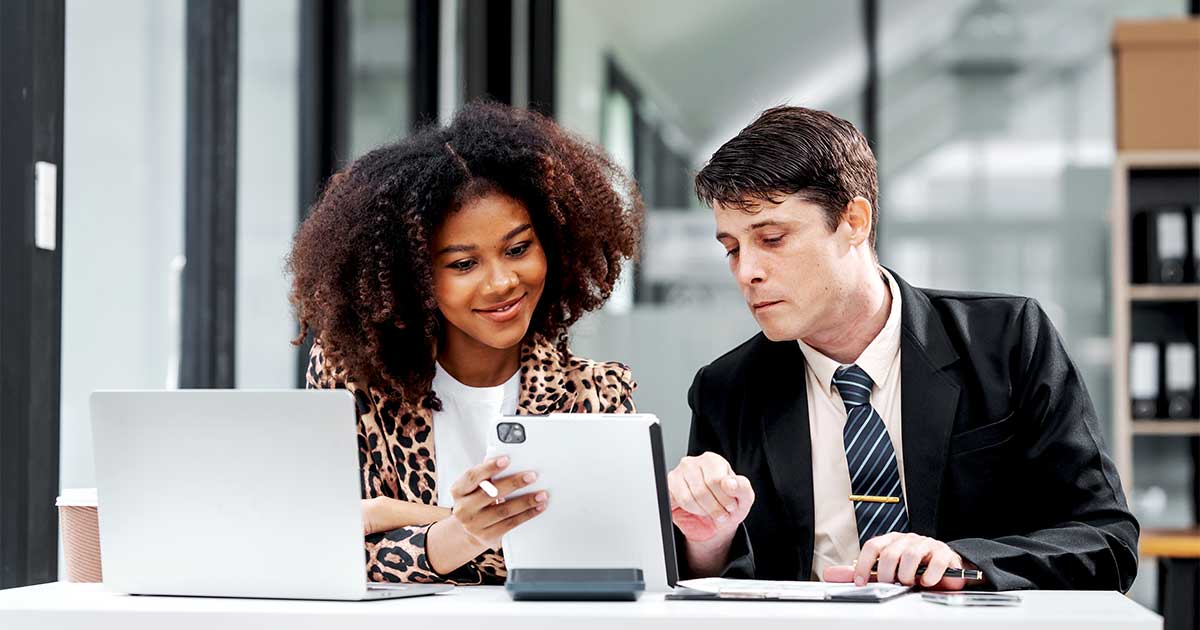Two colleagues review information on a tablet together at a desk with a laptop and notes.