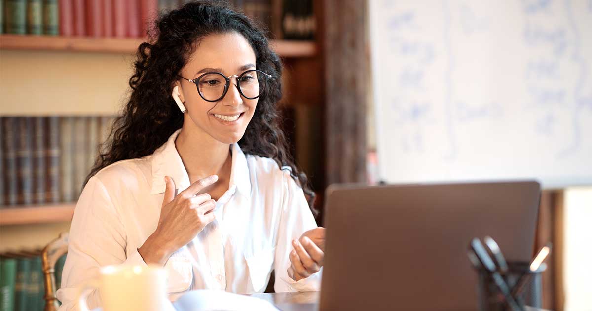 A smiling person in glasses and an earbud talks with someone on a video call at a laptop, with bookshelves behind them.