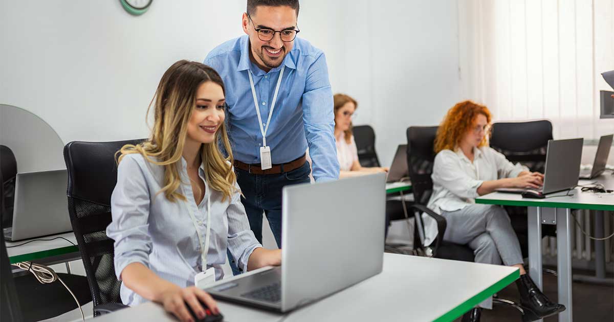 A colleague leans over to help someone at a laptop in a bright training room where others work at computers.